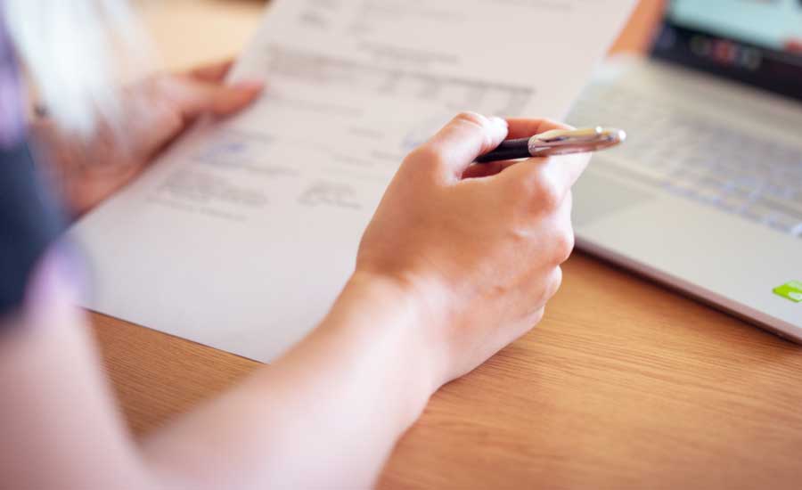 Mains tenant un stylo et écrivant sur un papier avec un ordinateur portable flou en arrière-plan