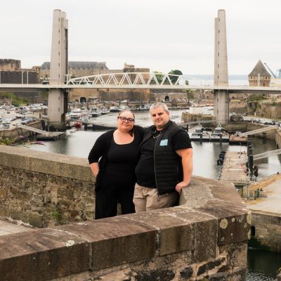 Charlotte & didier posant sur un mur de pierre devant le pont de Recouvrance et le port de Brest